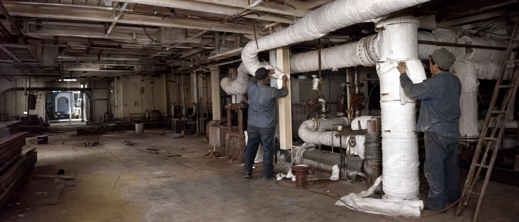 1960s shipyard engine room where workers wrap steam pipes in asbestos lagging, the kind of job that decides which asbestos trusts a claim can go to